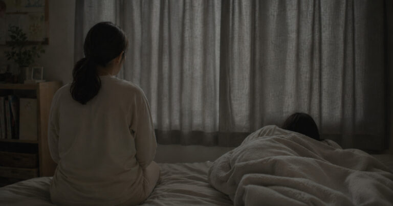 カーテンを閉めた薄暗い部屋で、布団にくるまる子どもとそばに座る母親の後ろ姿