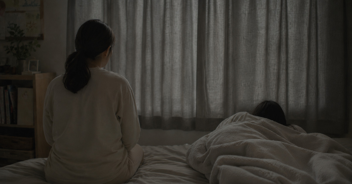 カーテンを閉めた薄暗い部屋で、布団にくるまる子どもとそばに座る母親の後ろ姿